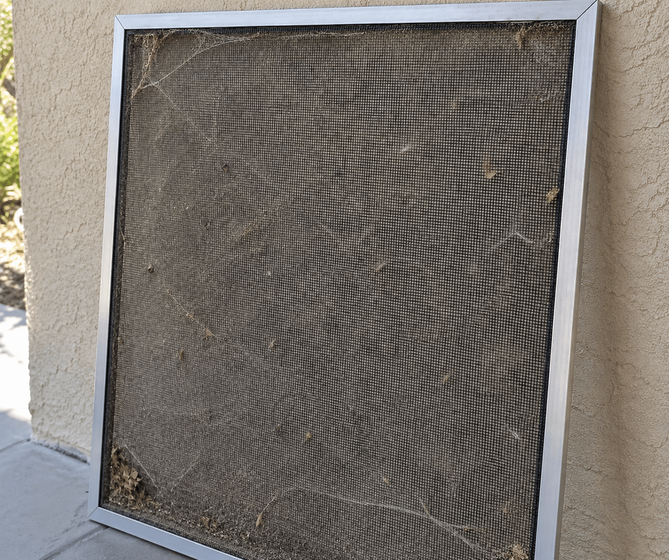 A window screen before cleaning, the mesh clogged with dust, cobwebs, and leaf debris